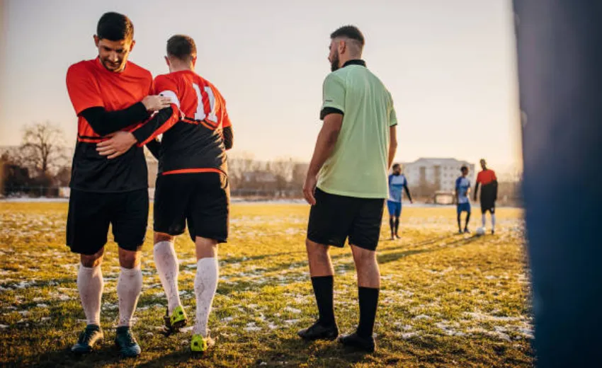 Due calciatori e un arbitro in piedi in un campo di calcio erboso, durante il in & out. Sono vestiti con abbigliamento e calzature sportive, è giorno con un cielo limpido sopra di loro.