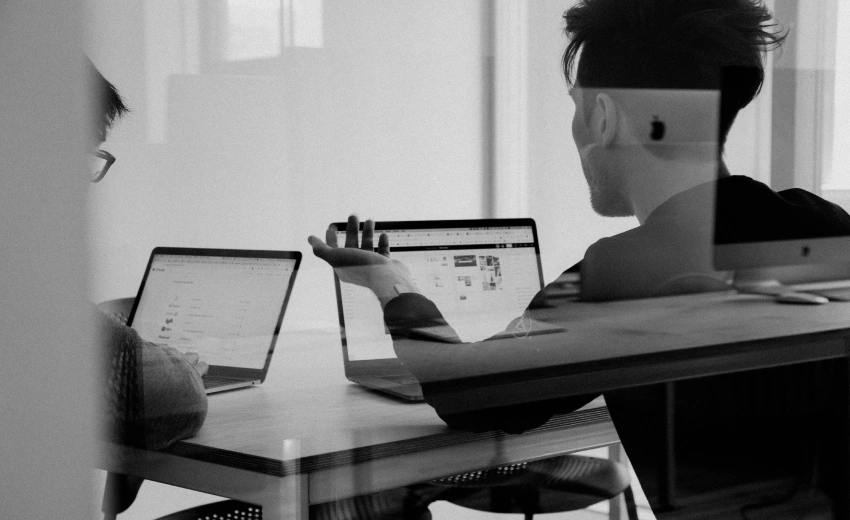 Due ragazzi in un ufficio di vetro guardano il computer e discutono le loro competenze utili alla azienda. L'immagine è in bianco e nero e si vedono dei riflessi sui vetri.