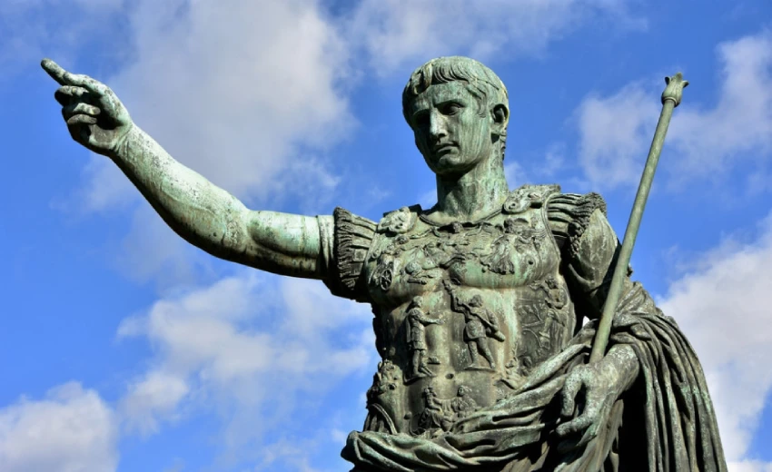 La statua dell'imperatore Augusto di colore verde rame con il braccio ad indicare l'azione, sapendo che corrisponde ad una reazione. Sullo sfondo il cielo azzurro con alcune nubi bianche.