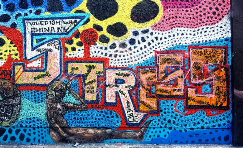 Un murales colorato con la scritta Stress e Fatica, vi è stilizzata in basso una donna seduta se si tiene la testa.
