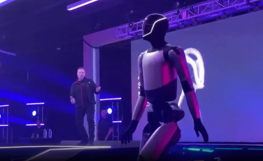 Visione di manager che vede un umanoide avanzato, presumibilmente l'Optimus di Tesla, in primo piano su un palco illuminato di viola e blu. L'umanoide ha un design elegante con sezioni nere e bianche e una visiera illuminata. Nello sfondo, Elon Musk, vestito con una giacca scura, sembra gesticolare o ballare. L'immagine cattura un momento di presentazione tecnologica.